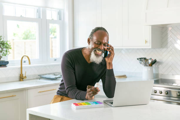 Senior black man speaking on the phone with his doctor about prescription medication Cheerful senior black man at home in the kitchen speaking on the phone with his doctor or pharmacist about prescription medication and daily supplements. The retired man is multi-tasking and has a laptop computer open on the kitchen counter. His daily pill box organizer containing his medication is next to the computer. Telemedicine, healthy lifestyle, and healthcare and medicine concepts concepts. pill prescription capsule prescription medicine stock pictures, royalty-free photos & images