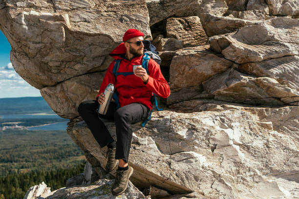 A male tourist is resting after a hard climb up the mountain. A tourist drinks coffee from a thermos in the mountains. A male traveler drinks hot tea from a thermos in the mountains. Copy space stock photo