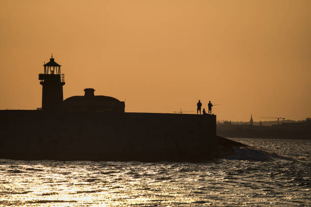 Telephoto evening view of silhouettes of people fishing at West Pier lighthouse seen from East Pier of Dun Laoghaire harbor, Dublin, Ireland stock photo