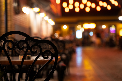 closeup rear view of black chairs with tables on restaurant terrace on a illuminated street at night with lights in the background
