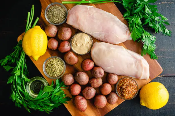 Raw chicken breasts, new potatoes, and other ingredients on a wood background