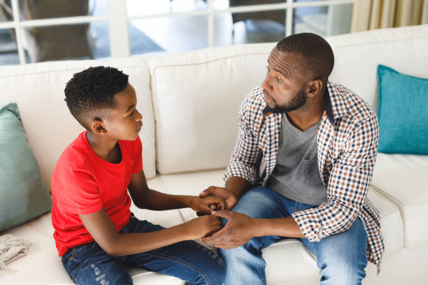 Serious african american father and son sitting on couch in living room talking and holding hands stock photo