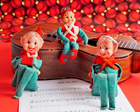 Christmas elves around an ukulele and joyous holiday music. Vintage elf figurines from the the early 1960s propped against a mahogany ukulele with Christmas holiday sheet music