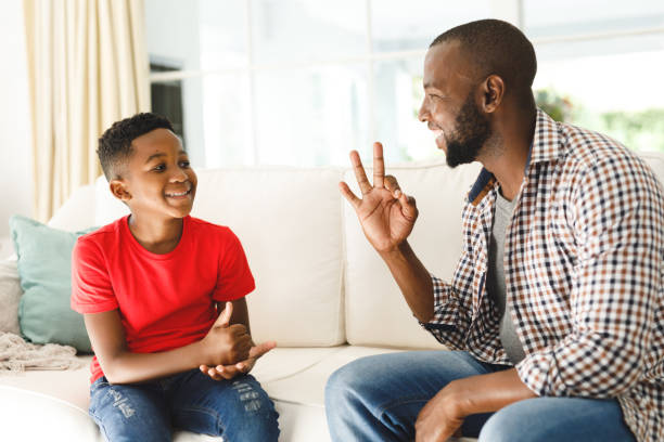 happy african american father with son sitting on couch in living room talking sign language - american sign language imagens e fotografias de stock