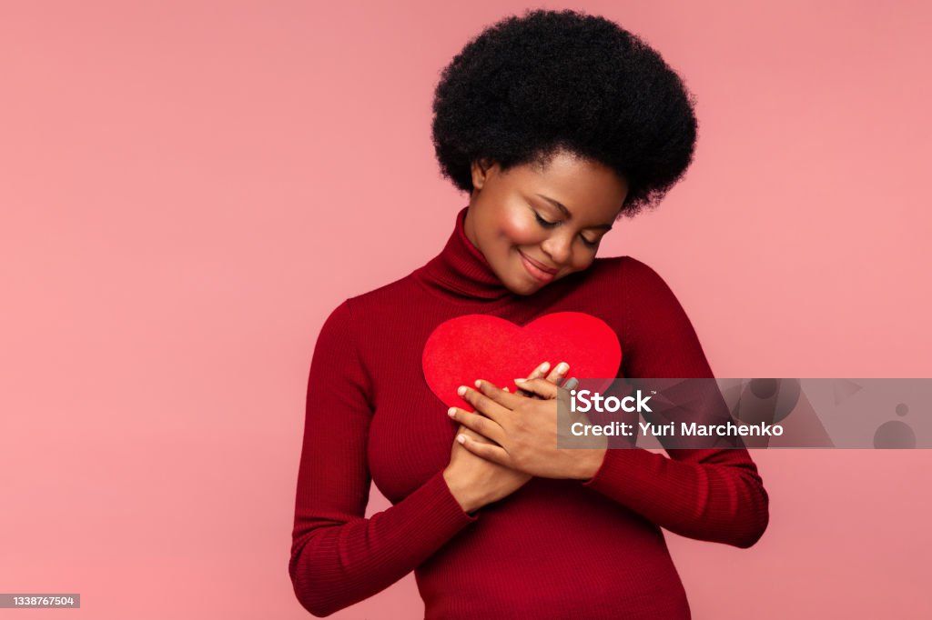 Happy african woman embracing red heart shaped postcard from lover or boyfriend on valentines day Valentine's day gifts and greetings: happy pleasured black woman holding red heart shaped postcard. African woman in love hugging romantic paper card from lover or boyfriend. Holiday sale for presents Heart Shape Stock Photo
