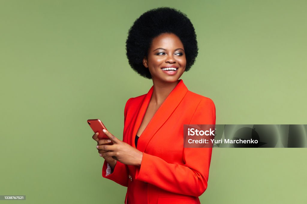 Successful african businesswoman with smartphone look aside with confident happy smile isolated Successful african businesswoman hold smartphone look aside with confident happy smile. Afro american young entrepreneur female in luxury red suit with mobile phone isolated over green studio wall Women Stock Photo