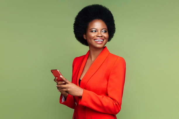Successful african businesswoman with smartphone look aside with confident happy smile isolated Successful african businesswoman hold smartphone look aside with confident happy smile. Afro american young entrepreneur female in luxury red suit with mobile phone isolated over green studio wall black business stock pictures, royalty-free photos & images