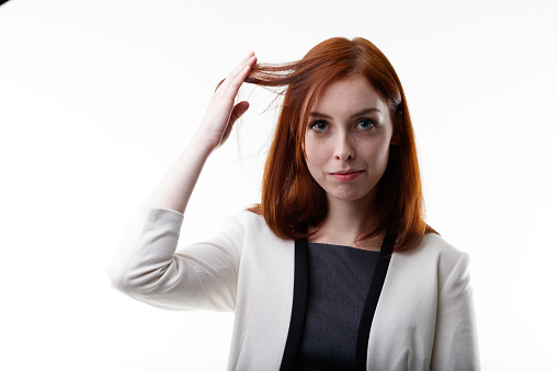 Serious young redhead woman holding up a strand of hair between her fingers as she looks at the camera in a head and shoulders portrait on white