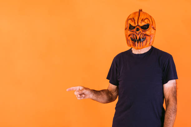 Man wearing terrifying pumpkin latex mask with blue t-shirt, pointing to the left side with his hands at the copy space, on an orange background. Halloween and days of the dead concept. stock photo