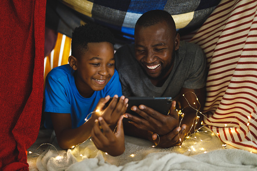 Happy african american father and son lying in blanket fort, using tablet. family spending time at home.