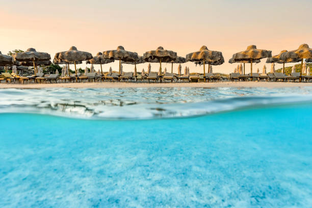 (Selective focus) Split shot, over under picture. Crystal clear, turquoise water and some beach umbrellas illuminated during a stunning sunrise. Grande Pevero Beach, Sardinia, Italy.