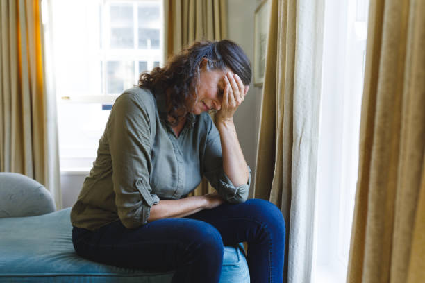 istock Stressed senior caucasian woman sitting on sofa by window holding head 1338803601
