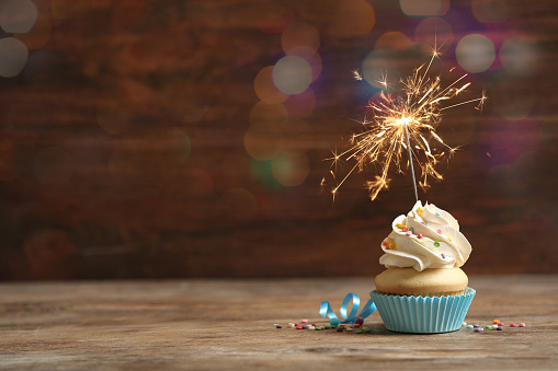 Birthday cupcake with sparkler on wooden table, space for text. Bokeh effect
