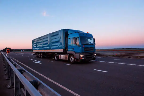 A long-distance truck driver is driving a tractor with a tilt semitrailer on an evening highway. Trucking business concept, indusrty