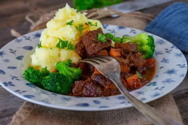 Homemade fresh cooked vanison stew or deer ragout served with mashed potatoes and vegetables on a plate with fork. Closeup and isolated view on rustic and wooden table background