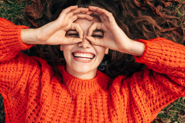 Top view of a happy woman wearing orange knitted sweater doing ok gesture like binoculars, smiling broadly and looking through fingers. Pretty girl pretend to looks through binoculars at something. stock photo