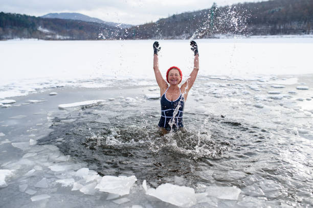 Front view of active senior woman in swimsuit splashing water outdoors in winter, cold therapy concept. A front view of active senior woman in swimsuit splashing water outdoors in winter, cold therapy concept. woman cold water swimming stock pictures, royalty-free photos & images