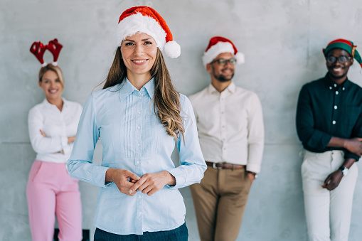 Shot of a confident smiling beautiful businesswoman with Santa Claus hat standing in front of her successful team in background. Multi-ethnic group of young business people with Santa Claus hats during Christmas party in the office.