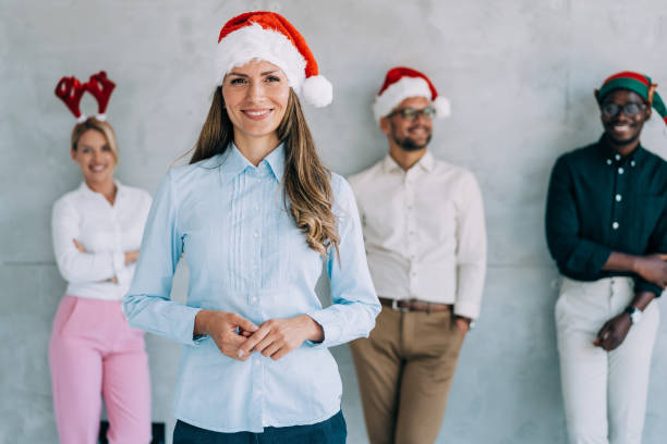 Business team leader. Shot of a confident smiling beautiful businesswoman with Santa Claus hat standing in front of her successful team in background. Multi-ethnic group of young business people with Santa Claus hats during Christmas party in the office. office christmas party stock pictures, royalty-free photos & images