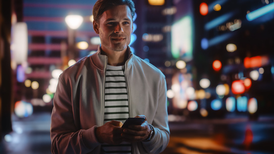 Handsome Man Using Smartphone Walking Through Night City Full of Neon Colors and Entertainment. Stylish Young Man Using Mobile Phone, Posting on Social Media, Online Shopping, Texting on Dating App