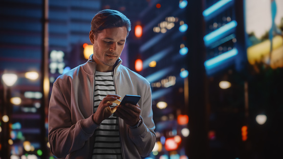 Hombre guapo usando teléfono inteligente caminando por la ciudad nocturna llena de colores de neón y entretenimiento. Joven con estilo usando un teléfono móvil, publicando en las redes sociales, compras en línea, mensajes de texto en la aplicación de photo