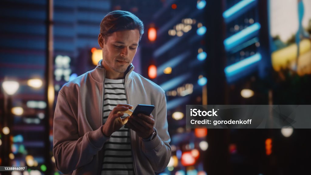 Handsome Man Using Smartphone Walking Through Night City Full of Neon Colors and Entertainment. Stylish Young Man Using Mobile Phone, Posting on Social Media, Online Shopping, Texting on Dating App Men Stock Photo