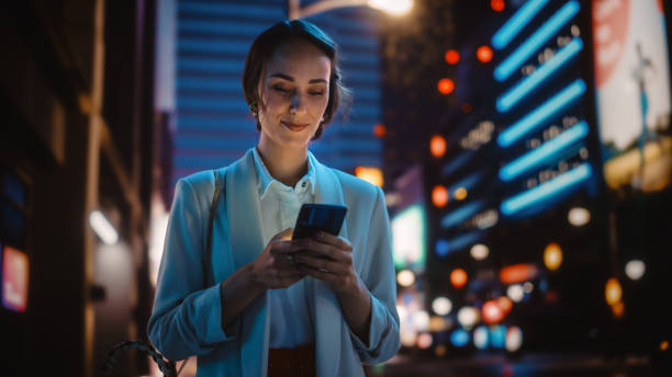 네온 빛으로 가득 한 밤 도시 거리를 걷는 스마트 폰을 사용하는 아름다운 젊은 여성. 휴대 전화를 사용하여 신중하게 여성 미소, 소셜 미디어 게시, 온라인 쇼핑, 문자 메시지. - mobile phone business technology elegance 뉴스 사진 이미지