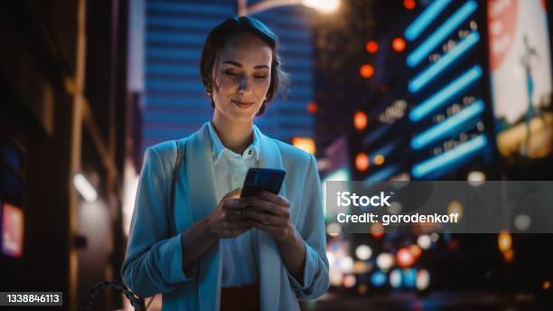 Beautiful Young Woman Using Smartphone Walking Through Night City Street Full Of Neon Light Smiling Thoughtfully Female Using Mobile Phone Posting Social Media Online Shopping Texting Stock Photo - Download Image Now