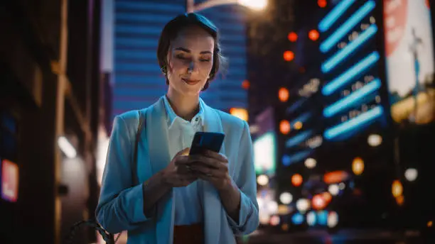 Photo of Beautiful Young Woman Using Smartphone Walking Through Night City Street Full of Neon Light. Smiling Thoughtfully Female Using Mobile Phone, Posting Social Media, Online Shopping, Texting.