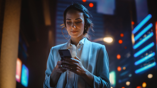 Hermosa joven con teléfono inteligente de pie en la calle Night City llena de luz de neón. Retrato de una hermosa mujer sonriente usando un teléfono móvil. photo