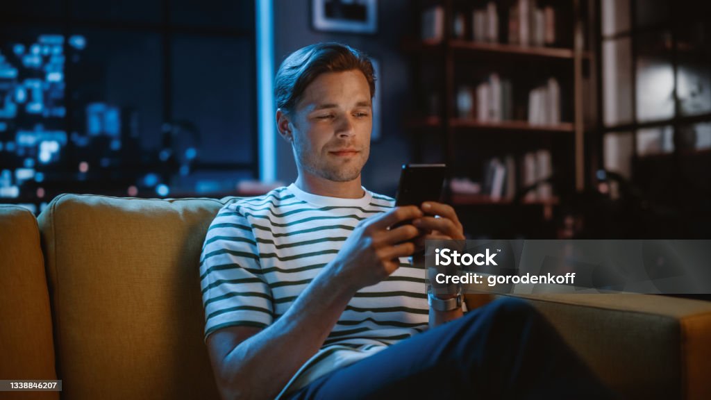 Handsome Caucasian Man Using Smartphone in Cozy Living Room at Home Sitting on a Sofa in the Evening. Doing Online Shopping, Browsing the Internet and Checking Videos on Social Media, Having Fun Mobile Phone Stock Photo