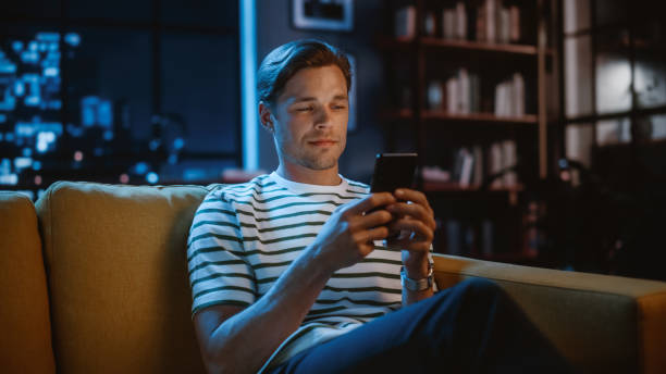 homem branco bonito usando smartphone em sala de estar aconchegante em casa sentado em um sofá à noite. fazendo compras online, navegando na internet e checando vídeos nas mídias sociais, se divertindo - beautiful caucasian one person romance - fotografias e filmes do acervo