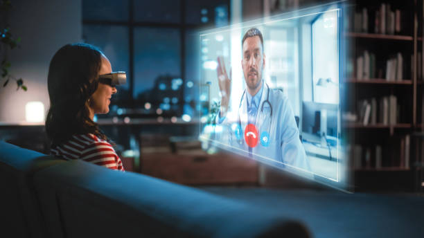 istock Young Woman Using Virtual Reality Headset At Home, Sitting on a Couch, Using VR Video Call Chat with Her Medical Consultant or Doctor. Sick Girl Calls the Doctor. Over the Shoulder 1338846213