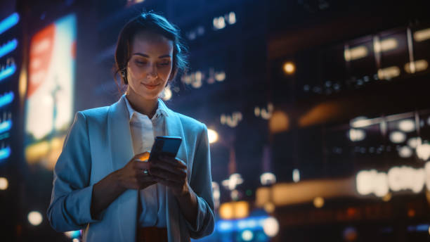 beautiful young woman using smartphone walking through night city street full of neon light. portrait of gorgeous smiling female using mobile phone. - woman on phone stockfoto's en -beelden
