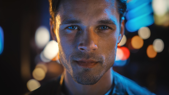 Portrait of Handsome Blonde Man Smiling and Looking at Camera, Standing in Night City with Bokeh Neon Street Lights in Background. Confident Young Man with Stylish Hair. Close-up Portrait.