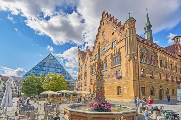 Ulm Ulm, Germany - August 28, 2021: People in front of the old town hall with the astronomical clock in Ulm. in the foreground the fish box, a historical fountain. In the background the glass pyrmid which contain the city library. thomas wells stock pictures, royalty-free photos & images