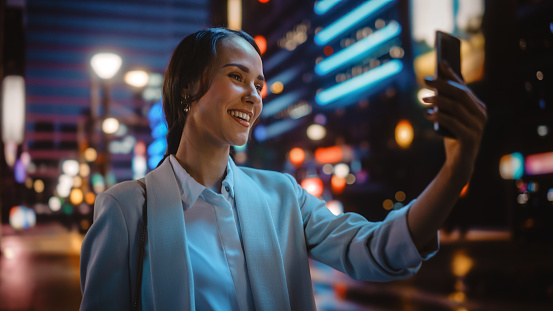 Beautiful Young Woman Using Smartphone for Video Call on Night City Street with Neon Lights. Portrait Smiling Female Using Mobile Phone Chatting with Family, Friends or Business Partners