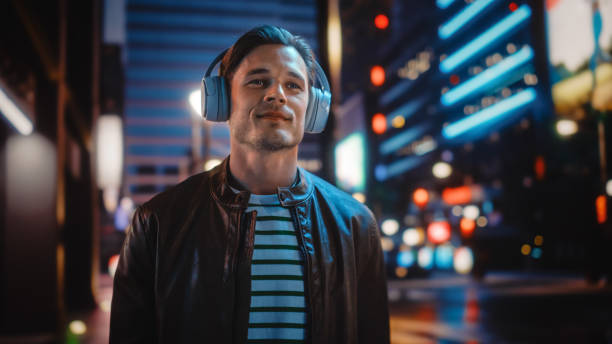 portrait d’un bel homme portant des écouteurs marchant dans la rue night city pleine de néons. smiling stylish man écoutant de la musique, podcast, talk-show. - men smiling headphones individuality photos et images de collection