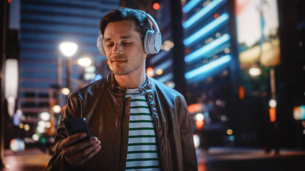 ritratto di bell'uomo che indossa le cuffie che cammina per night city street piena di luce al neon. uomo elegante sorridente che ascolta musica, si gode podcast, talk show. - men young adult human head blond hair foto e immagini stock