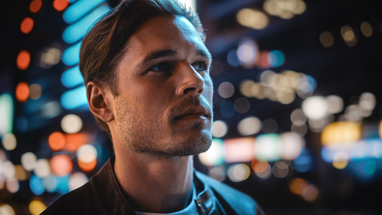 Portrait of Handsome Serious Man Standing, Looking Around Night City with Bokeh Neon Street Lights in Background. Focused Confident Young Man Thinking. Close-up Portrait Shot.