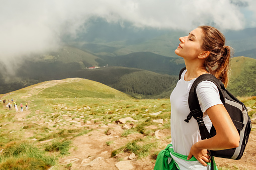 Woman hiker resting after climbing on mountain surrounded with clouds in Carpathians. Traveler with backpack breathing. Trip to summer Ukraine