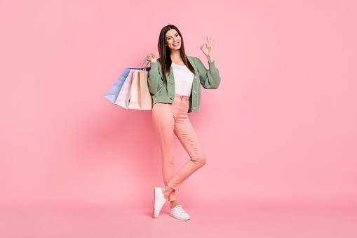 Full length photo of happy brunette young woman show okay sign smile hold bags isolated on pink color background.