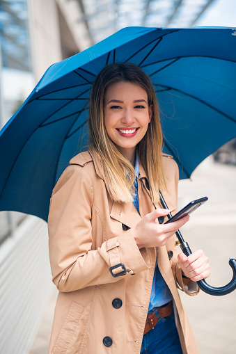 Portrait of a beautiful happy woman with blue umbrella and cellphone. She is looking art camera with a big smile on face