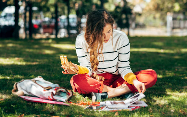 Horizontal image of a student female sitting on the green grass at the college campus on a sunny day, have lunch and studying. The hungry young woman takes a rest learning and eating in the park. stock photo