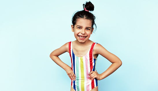 Joyful little girl wearing colorful swimwear has a toothy smile, enjoys her summer holiday with family, isolated over blue studio background with copy space for your text.
