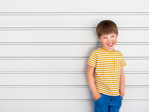 Portrait of smiling boy on white background with stripes. Cute kid stands hands in pockets near light grey wall. Child in yellow t-shirt and blue jeans laughs joyfully.