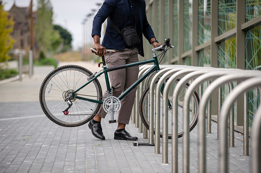 An unrecognisable mature black man wearing formal businesswear and a helmet on a summers day in a city. He is arriving at work and dismounting from his bicycle after sustainably commuting to work.