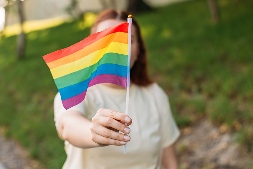Beautiful red lesbian woman with LGBT rainbow flag at sunset on green nature background. Happiness, freedom and love concept for same sex couples
