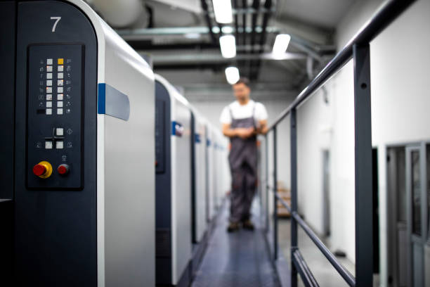 Close up view of modern offset printing press machine and operator in working uniform checking quality and controlling process of print.