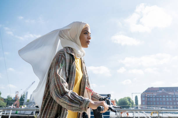 porträt einer jungen frau mit hijab auf e-scooter am flussufer in berlin - zurückhaltende kleidung stock-fotos und bilder